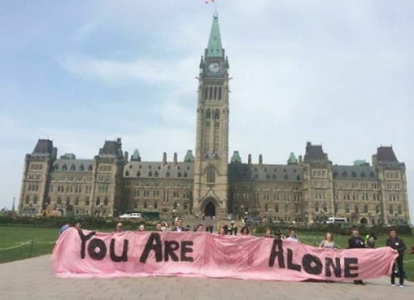 A large pink protest banner held in front of a government building reading you are alone.