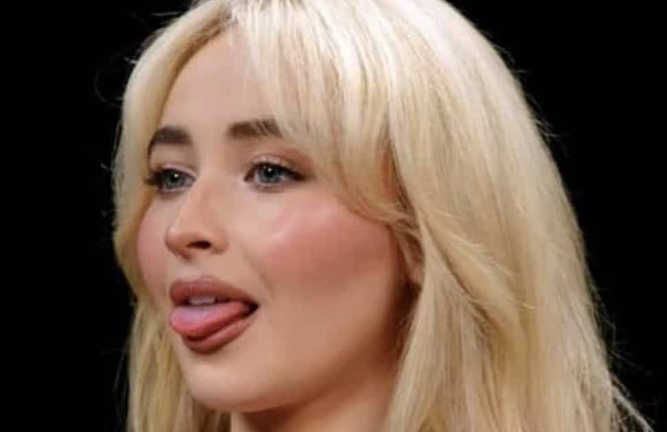 Close-up portrait of Sabrina Carpenter sticking her tongue out playfully on a black background.