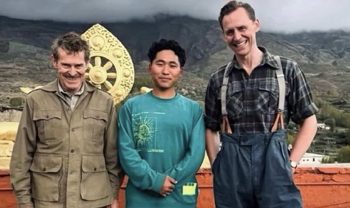 Willem Dafoe and Tom Hiddleston smiling alongside a local resident near scenic mountains.