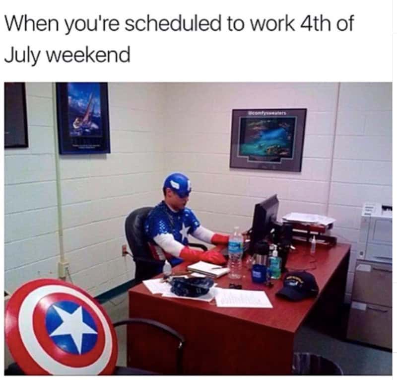 This relatable 4th of july meme targets holiday office workplace misery with a candid photo inside a sterile block-walled room. Under the caption, "When you're scheduled to work 4th of July weekend," an employee is seen sitting at a standard corporate desk typing on a computer while fully dressed in a realistic Captain America costume, complete with his vibranium shield resting nearby on the floor.