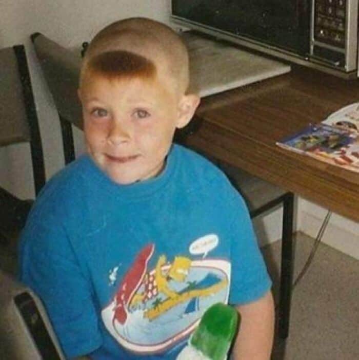 Redheaded boy holding a green popsicle with a completely shaved head except a tiny front fringe.