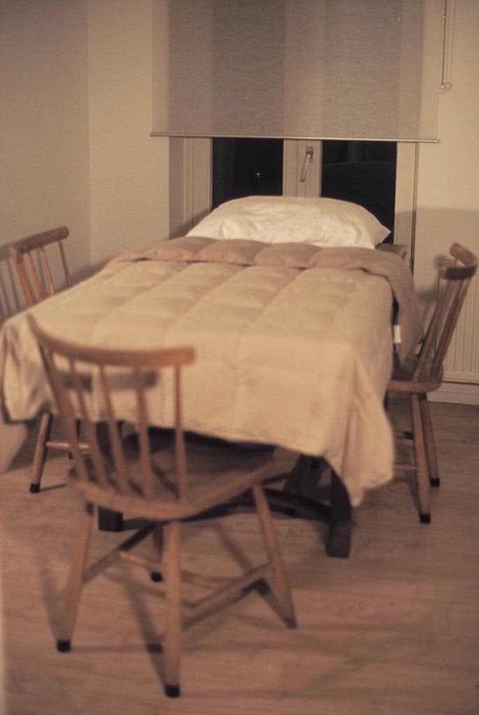 This terrifying airbnb meme presents a creepy, real-life photograph of a completely unhinged accommodation layout. Inside a dimly lit room, an ordinary wooden dining table surrounded by standard chairs has been converted into a bed, with a full-sized white pillow and a beige duvet comforter stretched directly across the tabletop under a window blind.