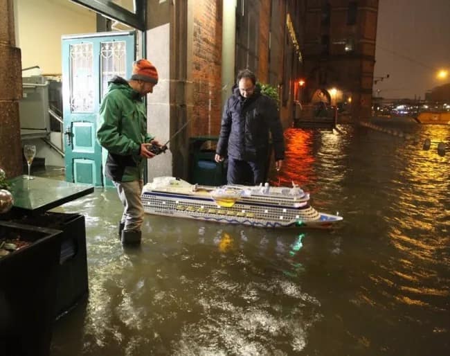 A spectacular entry in the best memes lineup shows two men taking full advantage of a severe urban weather crisis by navigating a highly detailed, illuminated remote-controlled miniature cruise ship down a deeply flooded city street.