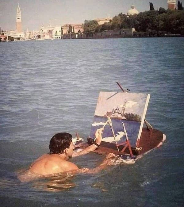 Shirtless man swimming in a lake while painting the Venetian cityscape on a floating easel.
