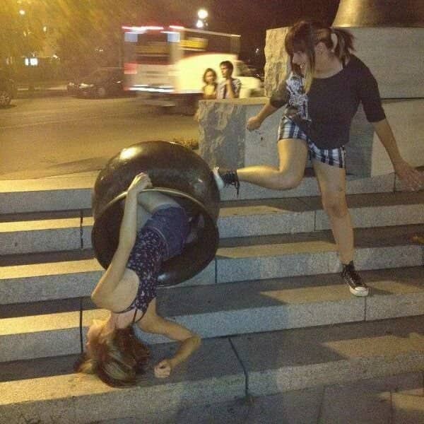 A girl rolling down stone stairs inside a large overturned trash bin while her friend watches.