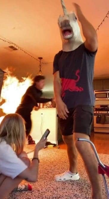 A chaotic living room scene with a kitchen fire while a man wears a unicorn mask.