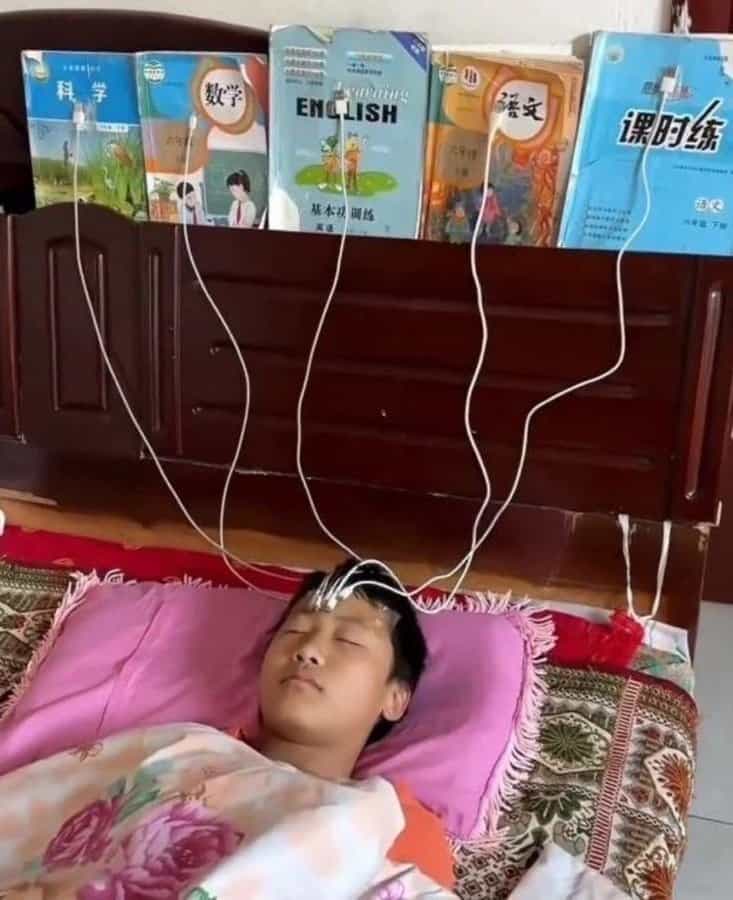 A child sleeping in bed with charging cables taped from school textbooks to their forehead.