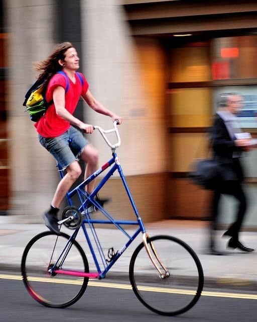 A long-haired cyclist riding a custom, ultra-tall blue bicycle frame down a city street.