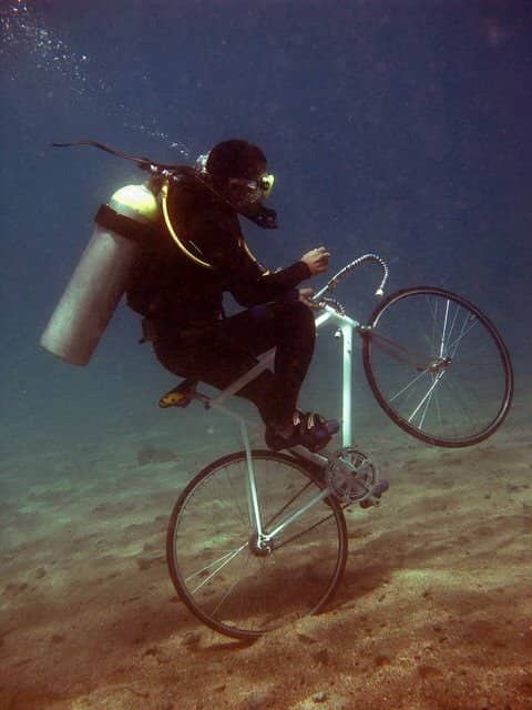 A scuba diver underwater riding a standard bicycle along the sandy ocean floor.