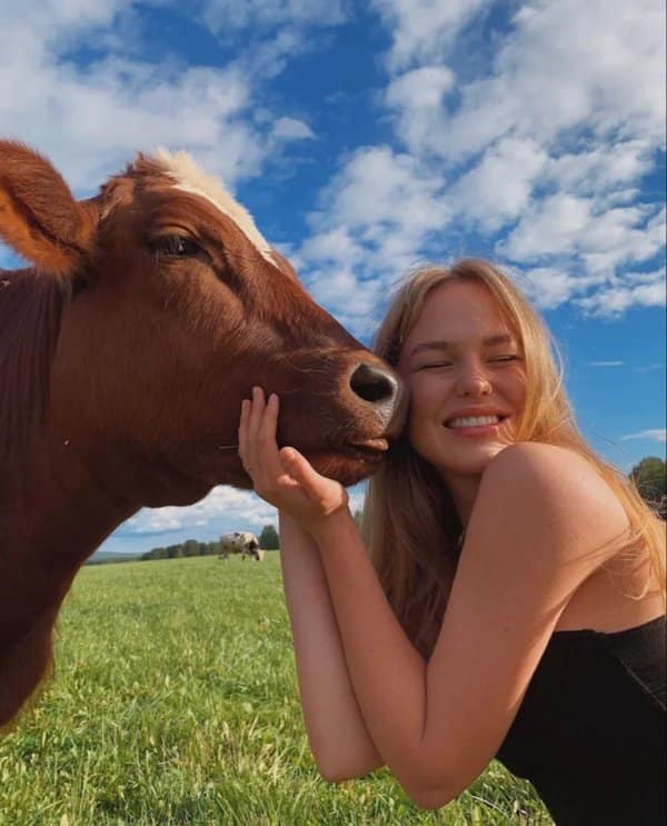 This exceptionally radiant cute photo captures a smiling blonde woman cuddling with a large brown dairy cow in a sprawling green pasture. Leaning her face against the cow's snout under a brilliant blue sky with fluffy white clouds, the woman evokes pure pastoral bliss.