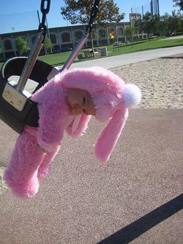 A delightfully silly cute photo showcases an infant dressed up in a fuzzy, bright pink bunny costume complete with long floppy ears and a white pom-pom. The baby hangs forward with a deadpan expression while sitting securely inside a black rubber playground bucket swing.