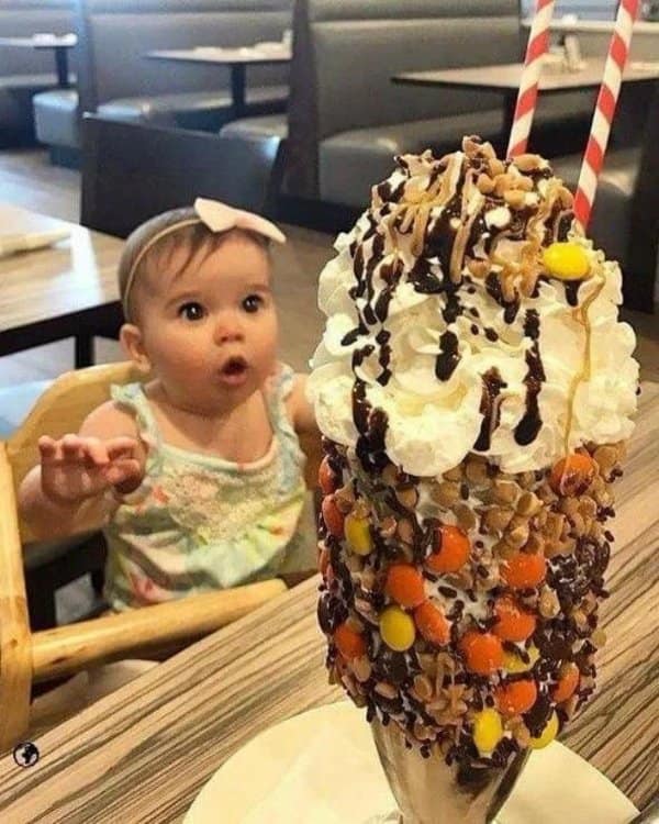 A hilarious and deeply cute photo highlights a baby girl sitting in a wooden restaurant high chair with her eyes wide and mouth open in pure astonishment. She looks in absolute awe at a giant, over-the-top gourmet milkshake piled high with whipped cream, hot fudge, and peanut butter candies.