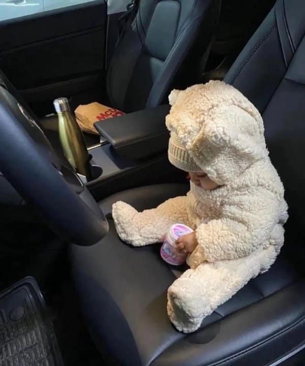 A comforting cute photo features a tiny baby dressed head-to-toe in a fluffy, cream-colored teddy bear fleece suit complete with little bear ears on the hood. The infant is sitting safely on the dark black leather passenger seat of a car, quietly playing with a small container.