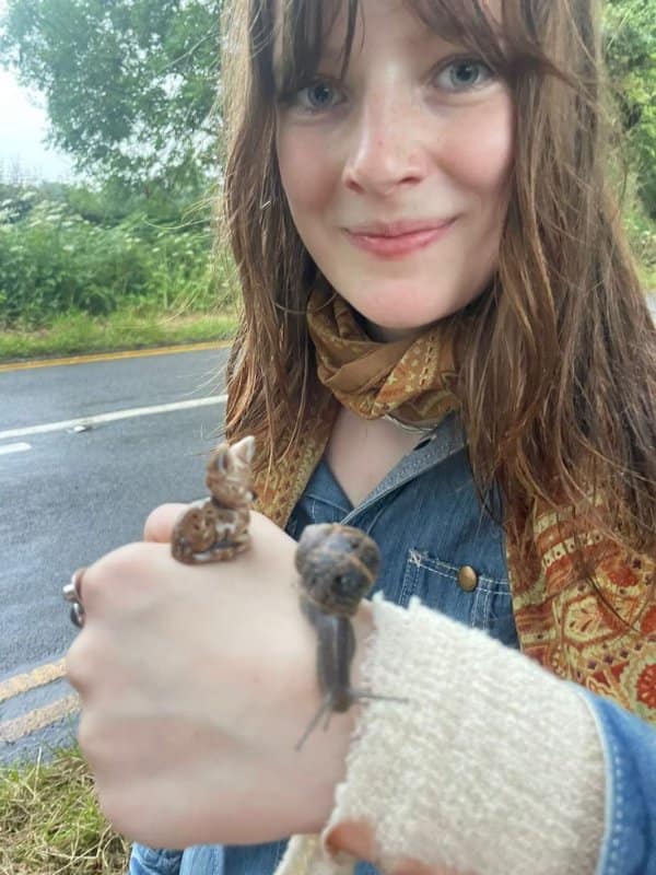 This charming and cute photo shows a smiling young woman holding her hand out toward the camera to display an unlikely pairing resting on her wrist: a tiny plastic deer figurine and a live garden snail slowly crawling along her sleeve cuff near a wet, paved countryside road.