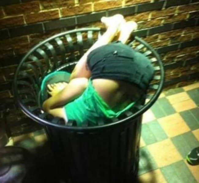 Person stuck upside down inside a metal public garbage can on a tiled sidewalk.