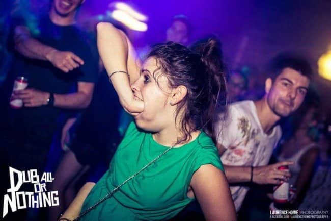 Woman on a purple-lit dancefloor shoving her entire fist inside her open mouth.