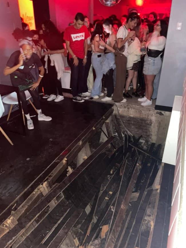 Group of clubgoers looking down in shock at a collapsed hole in the floorboards.