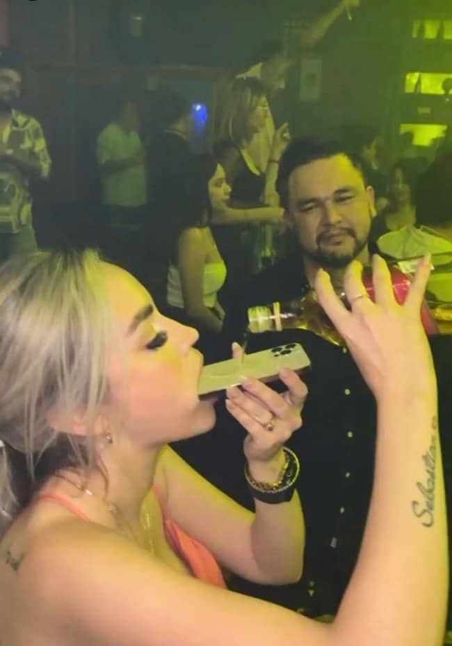 Woman drinking alcohol being poured flat down the screen of her phone at a club.