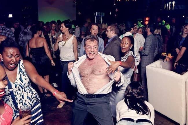 Middle-aged man wildly ripping open his button-down shirt on a crowded dance floor.