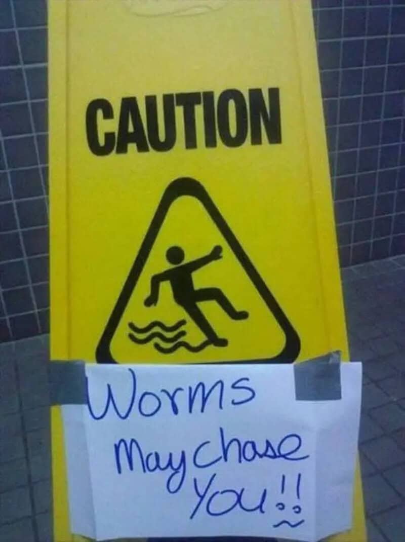 A chaotic public safety funny meme highlighting a standard yellow plastic "Caution" wet floor sign modified with a taped handwritten warning about being chased by rogue worms.