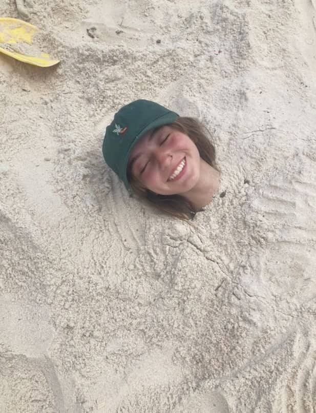A smiling girl wearing a green baseball hat buried up to her neck in sand.