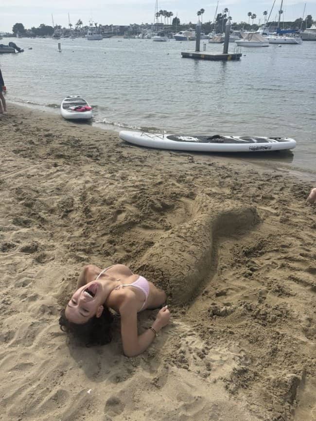 A laughing woman with a detailed sand sculpture mermaid tail lying on the beach.