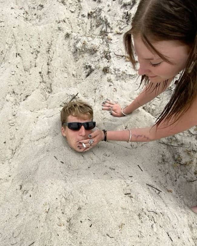 A woman putting a cigarette into the mouth of a guy buried in sand.