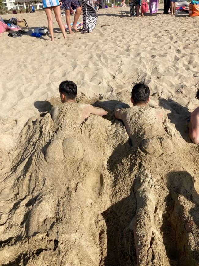 Two men buried in the beach with exaggerated glutes sculpted out of sand.