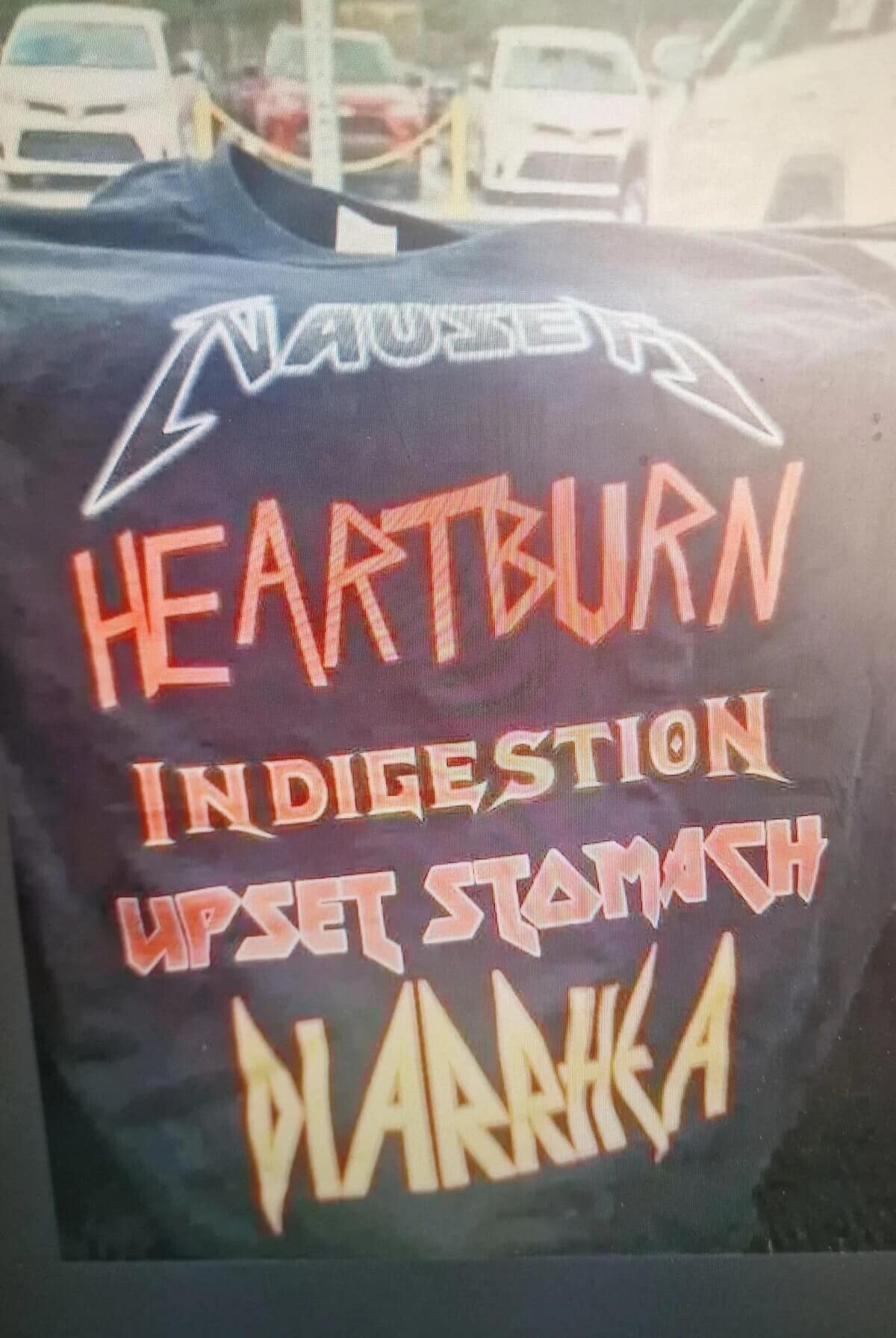 A custom apparel meme dump photograph showcases a black graphic t-shirt folded against a blurry parking lot background. The shirt features a parody logo styled like the heavy metal band Metallica that reads "NAUSEA," followed by a list of internal ailments printed in jagged metal fonts: "HEARTBURN INDIGESTION UPSET STOMACH DIARRHEA."