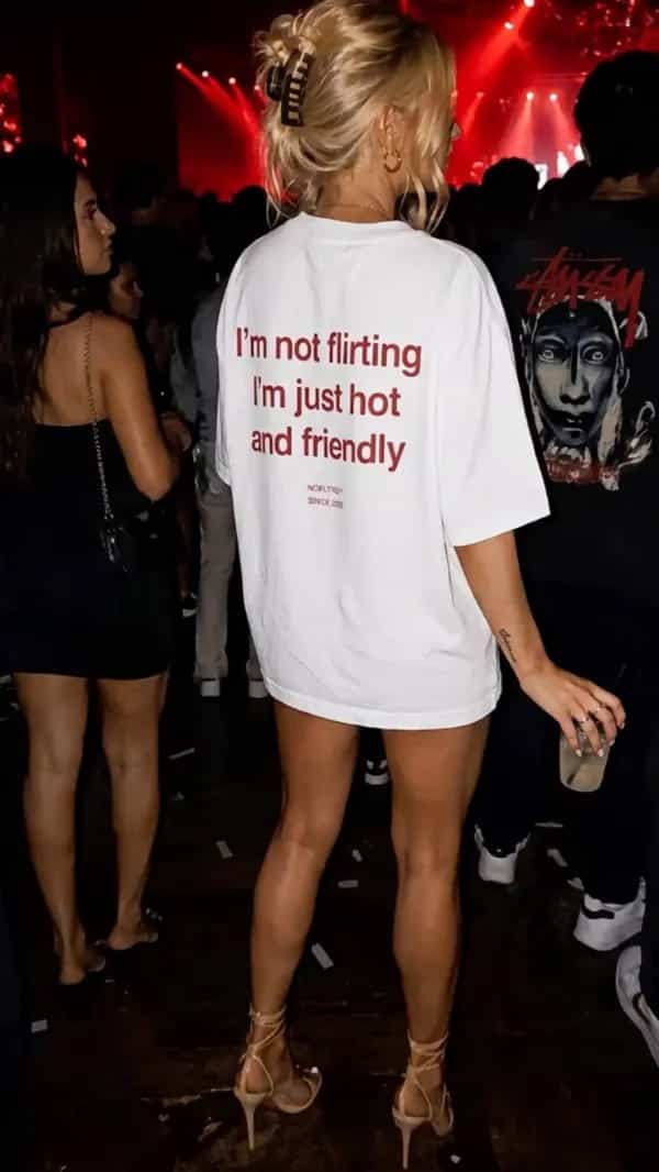 Blonde woman in party wearing white graphic t-shirt reading I'm not flirting I'm just hot and friendly.