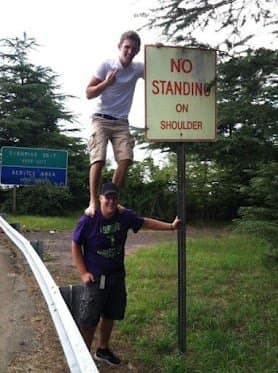 A literal interpretation road trip meme shows one guy standing directly on his friend's shoulders right next to a highway safety sign that forbids standing on the shoulder.