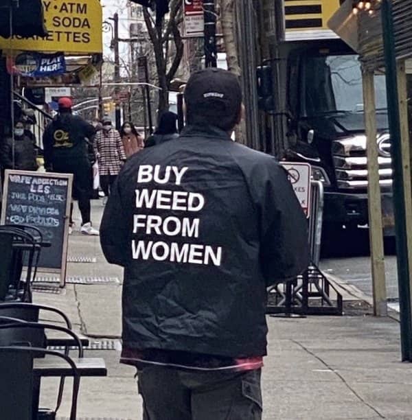 A man walking down a city street wearing a jacket that reads buy weed from women.